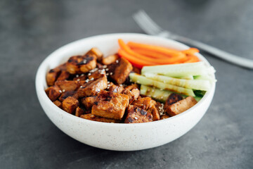 Bowl with roasted tempeh made of fermented soy beans, fresh vegetable sticks with sauce and sesame seeds on the table. Plant based protein. Healthy cooking and eating. Go vegan. selective focus