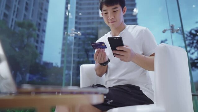 Asian man making online reservation with credit card, secure transactions, using smartphone and laptop, online shopping verification, entering ccv number, indoor shot