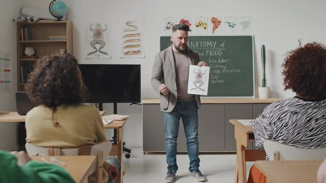 Tracking in of male Caucasian teacher holding image of frog anatomy, teaching high school students, standing by blackboard in classroom at daytime