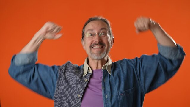 Portrait of excited jubilant overjoyed elderly bearded man 50s 60s doing winner gesture celebrate clenching fists say yes isolated on solid orange background studio
