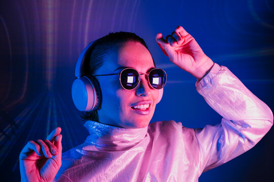 Portrait of a smiling dancing woman in sunglasses and headphones in neon light. Music lover. Silent disco. Woman in holographic clothes on the background of music vibes background. Futuristic Party.