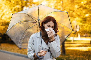 Sad young european female in raincoat with umbrella blows her nose in napkin, suffer from ill and...