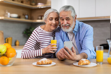 Portrait Of Happy Senior Spouses Using Smartphone During Breakfast In Kitchen