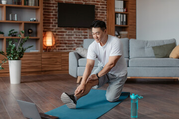 Active asian mature man in sportswear stretching leg muscles in front of laptop, watching online tutorials