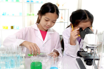 Adorable Asian schoolgirl in lab coat pouring green reagent solution from test tube into beaker while another girl looking through microscope, kids do science experiment, happy smiling young scientist