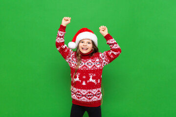 A little girl in a red sweater with reindeer and a Santa Claus hat waiting for the holiday on a green isolated background. The concept of Christmas.