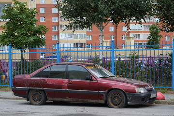 An old broken rusty abandoned dark red car stands near the blue fence, Soyuzny Prospekt, St. Petersburg, Russia, September 2022