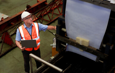 A male engineer wearing a safety suit and a hard hat uses a laptop to inspect machine maintenance in a factory.