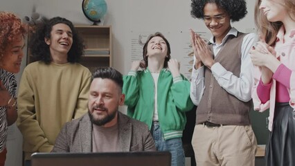 Low angle of multiethnic high school students clapping hands, standing around male teacher who sitting at desk in classroom, checking exam results on laptop computer in afternoon - Powered by Adobe