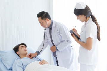 Senior doctor and young male patient who lie on the bed while checking symptom, consult and explain with nurse taking note and supporting in hospital wards.