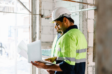 Two construction engineers in vest with wear safety hardhat hold blue print and laptop computer and talk about construction work on building site