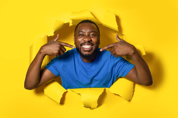 Shot of smiling african american man standing in torn paper hole, pointing at his perfect smile and...