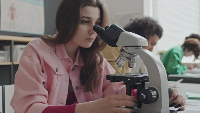 Chest-up of long-haired Caucasian girl using microscope, writing in notebook, sitting at desk next to Biracial guy who dissecting frog on biology lesson in afternoon