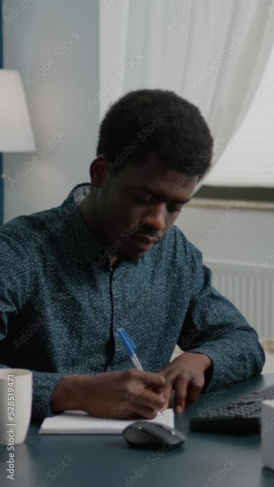 Poster vertical video: black african american freelancer taking notes on notepad and looking into computer,