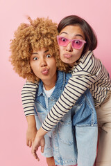 Lovely multiethnic women stand closely to each other keep lips rounded look surprisingly at camera have good relationships isolated over pink background. Friendship and togetherness concept.