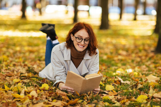 Smiling smart pretty young european woman in glasses and raincoat reads book, lies on yellow leaves in park