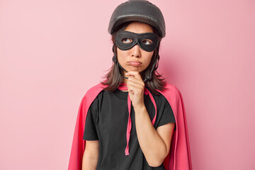 Sad brunette Asian woman holds chin looks unhappily aside being deep in thoughts dressed in superhero costume with cloak thinks about future isolated over pink background makes plans for masquerade