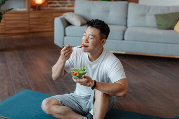 Healthy lifestyle concept. Relaxed asian man eating fresh vegetable salad, sitting on yoga mat...