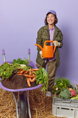 Hard working busy female gardener holds watering can dressed in workwear collects seasonal harvest happy to grow big vegetables in own garden looks happily away. Gardening and agronomy concept