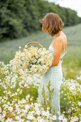 Young stylish woman in dress enjoys summer time, spending leisure time, gathering wild flowers in daisy field