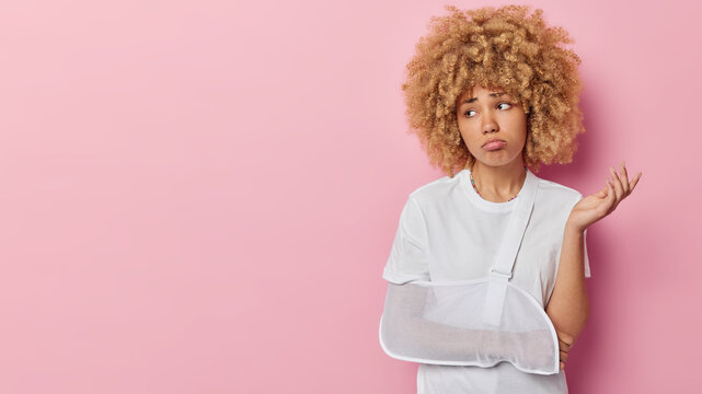Puzzled displeased curly haired woman has broken arm after workout or accident wears arm sling feels confused and upset feels pain going to visit doctor focused aside isolated over pink background