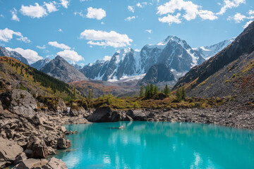 Pure turquoise alpine lake with view to forest hill and giant snow mountains in autumn sunny day. Glacial lake against huge snow covered mountain range in bright sun. Vivid colors in high mountains.