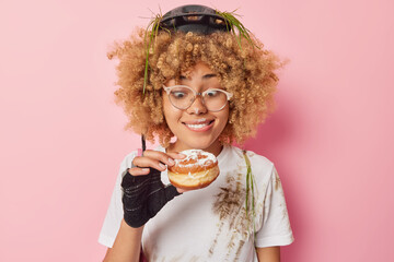 Horizontal shot of curly haired woman bites lips looks at appetizing doughnut wants to eat wears...