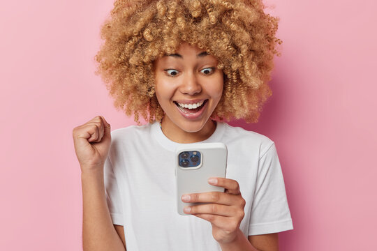 Cheerful curly haired young woman holds smartphone clenches fist celebrates success triumphs over something reads something awesome glad to achieve bonus in app dressed in casual white t shirt