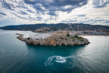 Fotografía aérea del castillo de Peñíscola con el cielo nublado y algunas motos de agua.