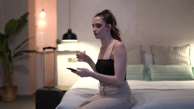 Young beautiful woman drinking a glass of water and scrolling through her phone on her bed