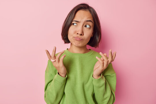 Clueless puzzled Asian woman shrugs shoulders feels hesitant about something spreads hands sideways feels unaware dressed in casual green pullover isolated over pink background. So what to do