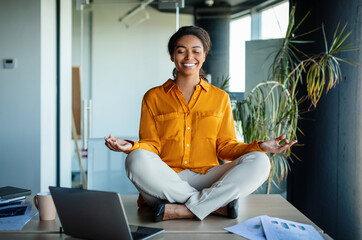Office zen. Calm black businesswoman meditating with closed eyes on office desk, sitting at...