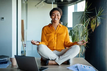 Tuinposter Office zen. Calm black businesswoman meditating with closed eyes on office desk, sitting at workplace in lotus position © Prostock-studio