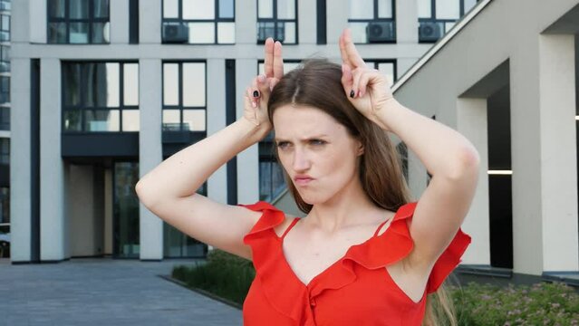 Aggressive bully woman with long hair wearing red elegant dress holding fingers above head showing horns, arrogant and stubborn, ready to attack. Outdoor shot with modern building on background.