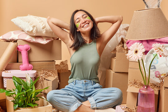 Positive Asian woman keeps hands behind head smiles gently sits crossed legs on floor dressed in t shirt and jeans happy to buy new apartment relocates to other place of living poses in messy room