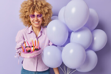 Horizontal shot of positive woman celebrates special occasion poses against purple background with bunch of inflated balloons and sweet festive cake prepares for special occasion. Holiday concept