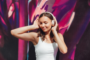 A young happy  woman dressed in a white top and purple skirt, headphones, listening to music, dancing on a city street near the wall of a building. The concept of the lifestyle of urban youth People.