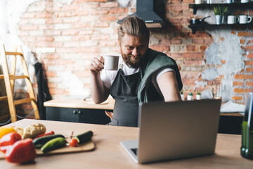 Smiling bearded chef browsing laptop at counter in kitchen