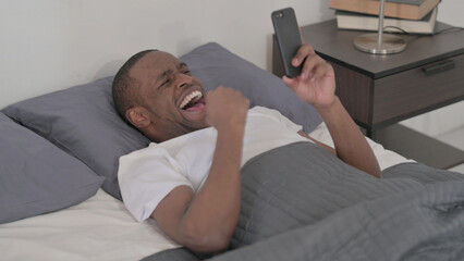 Fototapeta premium African Man Celebrating Success on Smartphone in Bed