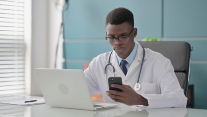 Fototapeta na wymiar Young African Doctor using Smartphone while using Laptop in Office