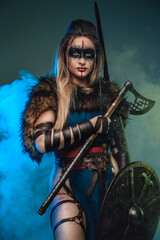 Nordic huntress woman with strup back longbow holding shield and axe against teal background with smoke. - obrazy, fototapety, plakaty