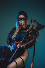 Shot of valkyrie with makeup and fur coat holding shield and hatchet against turquoise background.