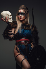Shot of medieval female hunter with fur holding shield and skull head against gray background.