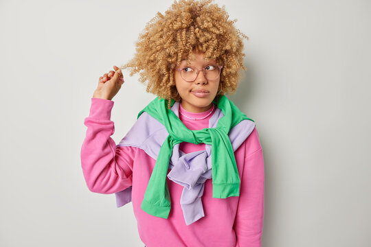 Photo of lovely woman curls hair being deep in thoughts concentrated away considers something wears spectacles casual pink jumper pullovers tied over shoulders isolated over grey background.