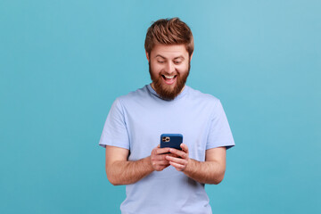 Portrait of happy excited bearded man using online service on mobile phone and expressing pleasant emotions, texting messages for friends. Indoor studio shot isolated on blue background.