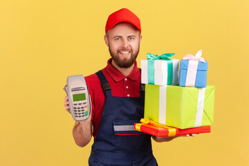 Portrait of positive delighted courier man wearing blue overalls holding stack of present boxes and pos terminal for online paying. Indoor studio shot isolated on yellow background.
