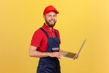 Side view of worker man wearing blue overalls, red T-shirt and cap working on laptop, expressing positive emotions, looking at camera. Indoor studio shot isolated on yellow background.