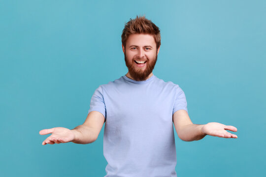 Portrait Of Pleased Bearded Man Sharing Opening Hands Looking At Camera With Kind Smile, Greeting And Regaling, Happy Glad To See You. Indoor Studio Shot Isolated On Blue Background.