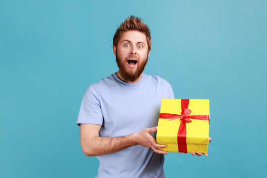 Portrait of excited handsome bearded man holding yellow present box with red ribbon, looking at camera with amazement, congratulating . Indoor studio shot isolated on blue background.