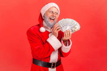 Fototapeta na wymiar Portrait of elderly man with gray beard wearing santa claus costume with excited expression, greedy male with fan of dollars, looking at banknotes. Indoor studio shot isolated on red background.
